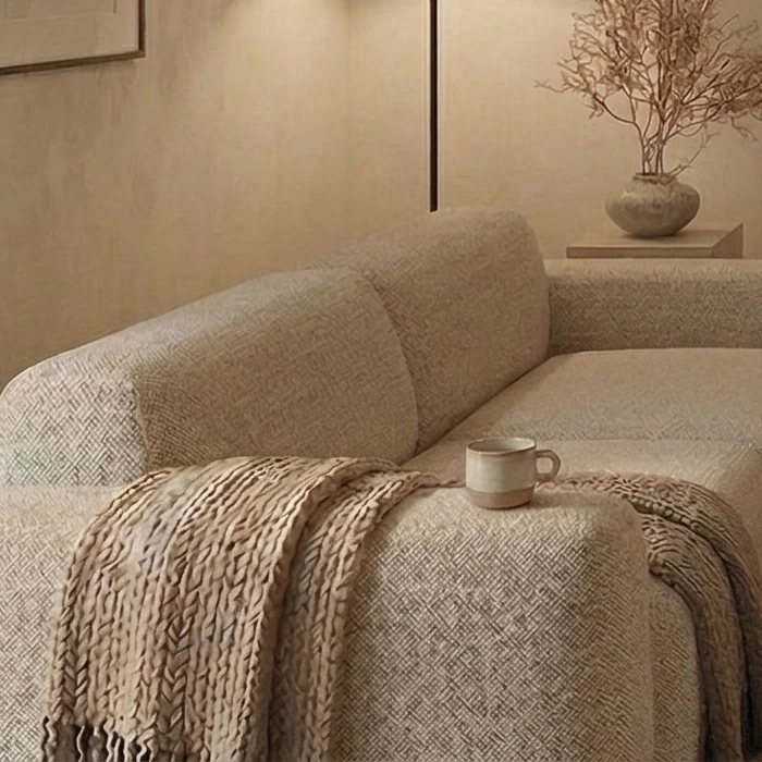 Chunky knitted throw on a sofa with textured upholstery, a ceramic mug beside it and a dried branch in a ceramic vase in the background