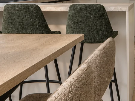 Olive-green bar stools with textured upholstery and black metal legs at the peninsula, a white end panel running down to the floor on the right, and the dining table top and a beige upholstered chair in the foreground