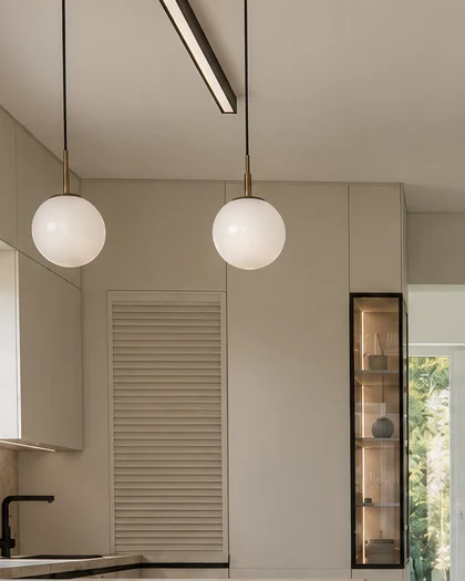 Two white globe lights above the peninsula, with a horizontal slatted front and a narrow, black-framed glass display cabinet behind them and the black linear light above