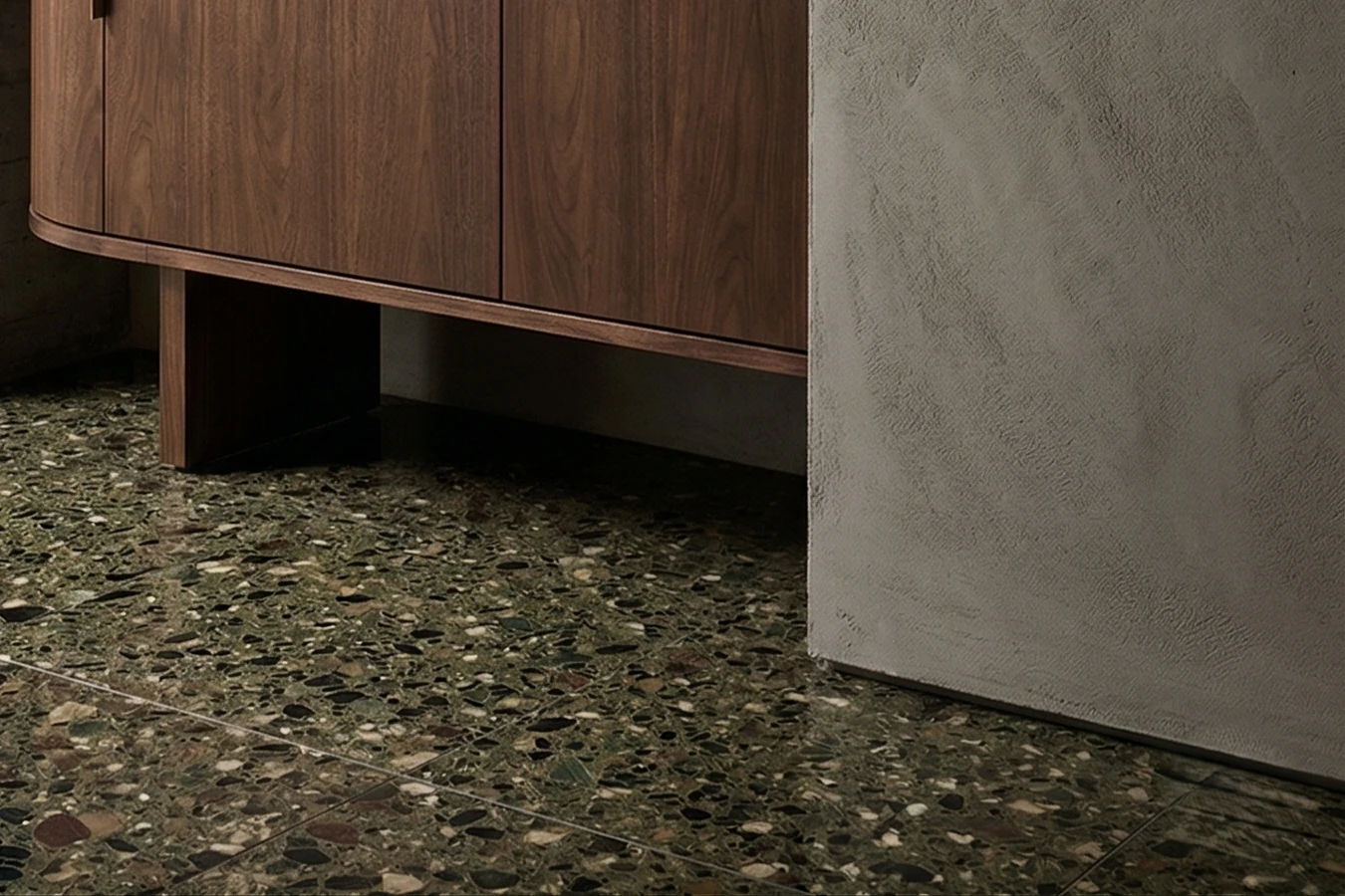 Dark green terrazzo floor with larger chips, the base of a walnut sideboard and a wall corner with a textured finish